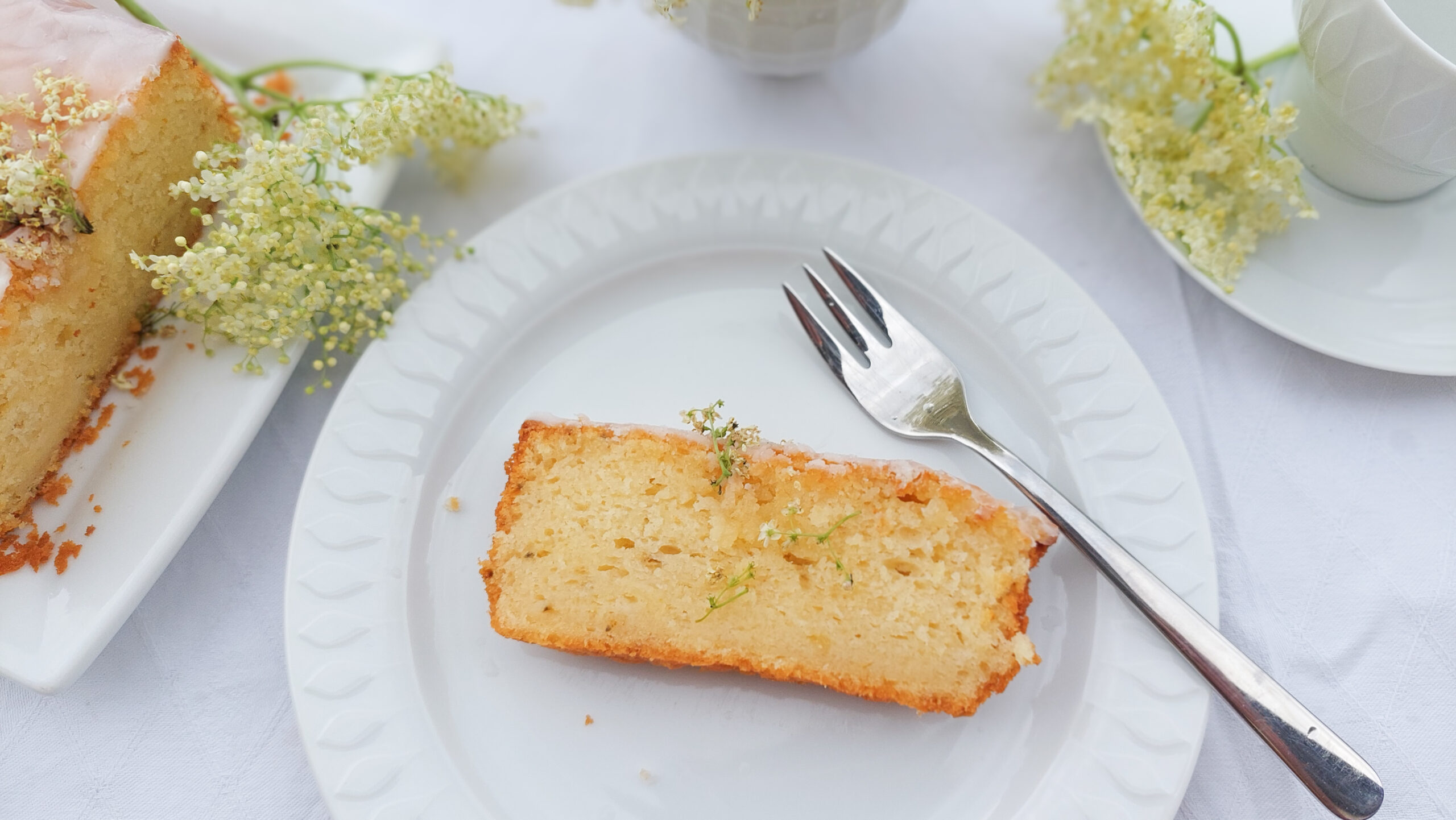 Elderflower cake IV Piece of elderflower cake on a plate, cake and coffee cup in the background