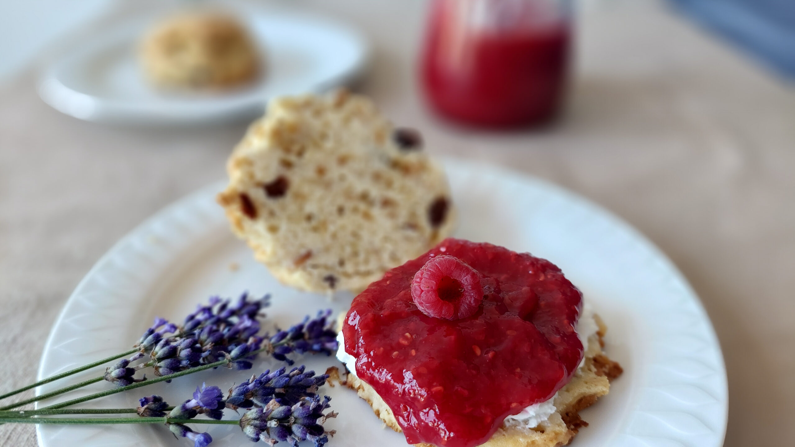 Scone with raspberry lavender jam, a glass of jam in the background and lavender stems next to the scone on the plate