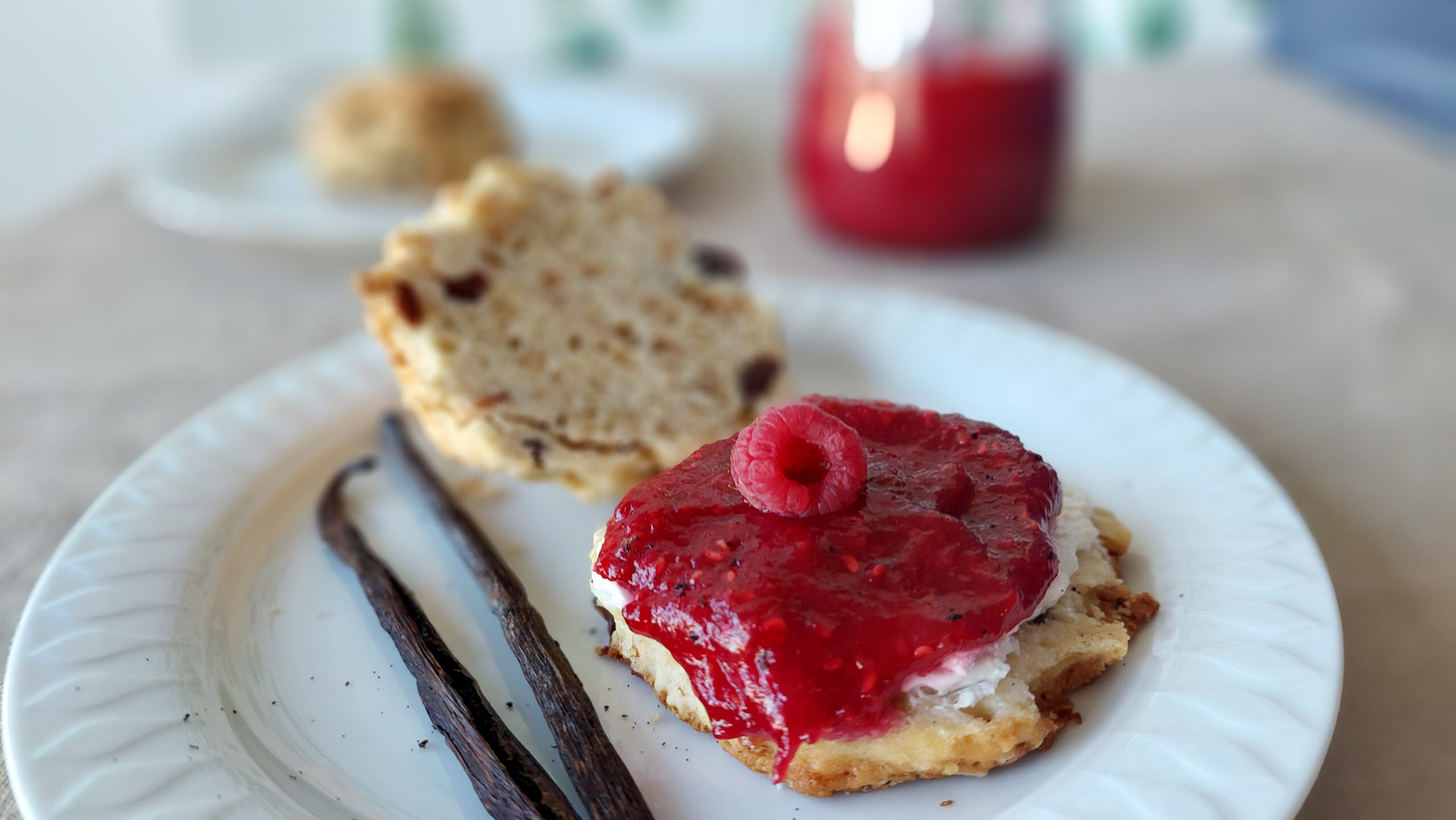 Scone with raspberry vanilla jam, a glass of jam in the background and vanilla pods next to the scone on the plate