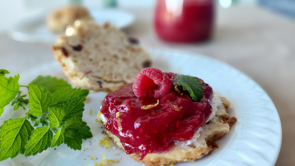 Scone with raspberry jam with lemon and lemon balm, decorated with a raspberry, lemon peel and lemon balm leaves; a sprig of lemon balm next to the scone and a jar of jam in the background