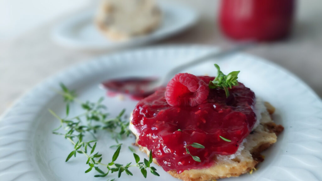 Raspberry jam on a scone, decorated with basil, sprigs of thyme lying next to it, a jar of jam in the background