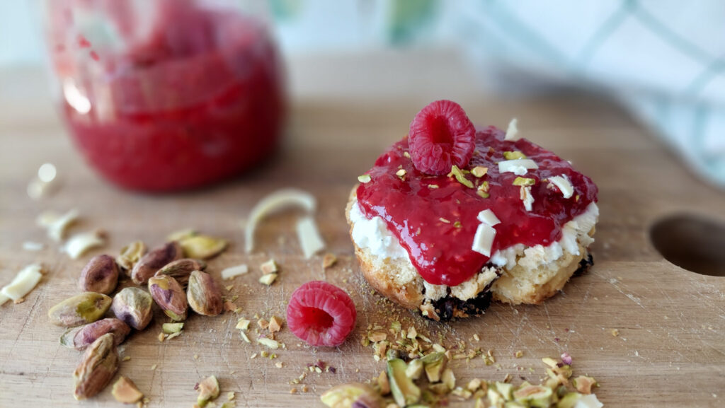 scone with raspberry jam on a wooden board; the scone is decorated with white chocolate curls and chopped pistachios, a jam jar in the background