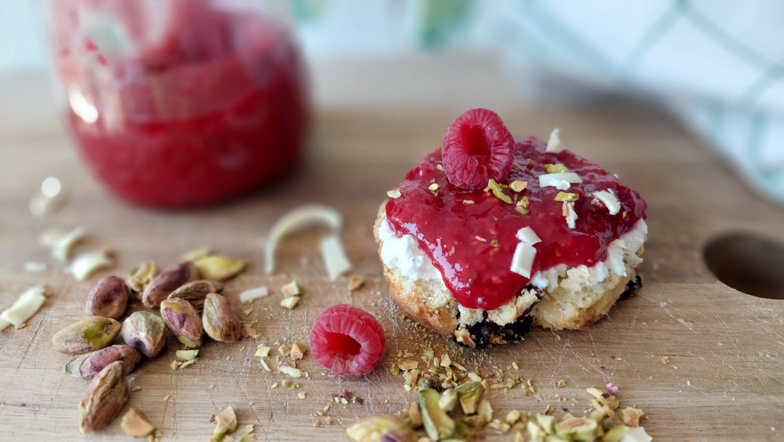 scone with raspberry jam on a wooden board; the scone is decorated with white chocolate curls and chopped pistachios, a jam jar in the background
